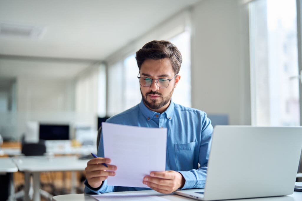 Young busy latin professional business man checking document working at laptop computer in office. Serious businessman accountant expert reading legal paper company file overview at workplace.