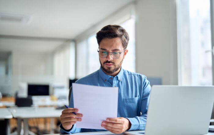Young busy latin professional business man checking document working at laptop computer in office. Serious businessman accountant expert reading legal paper company file overview at workplace.